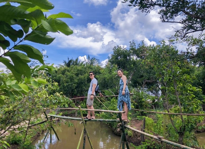 Two people standing on a bamboo bridge over a water body, surrounded by lush greenery. 2