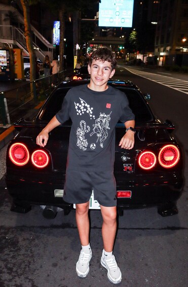 A person standing next to a black sports car with visible rear lights and a street sign in the background. 3