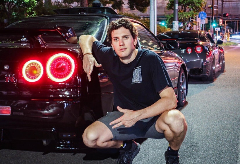 A person crouching next to a black sports car with its rear lights on, in an urban street at night. 2