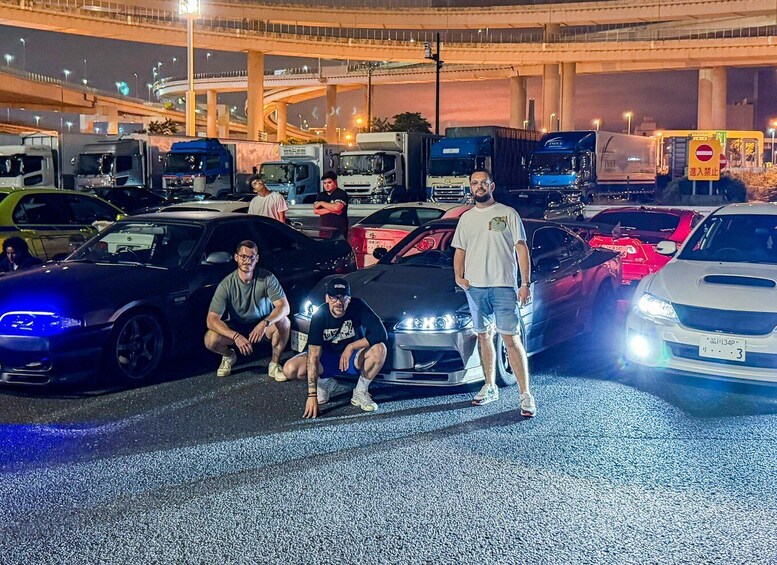 A group of men posing with sports cars in a parking lot at night. 3