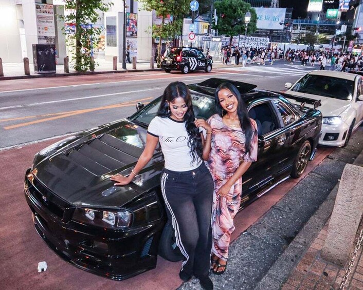 Two women posing with a black sports car on a city street at night. 2