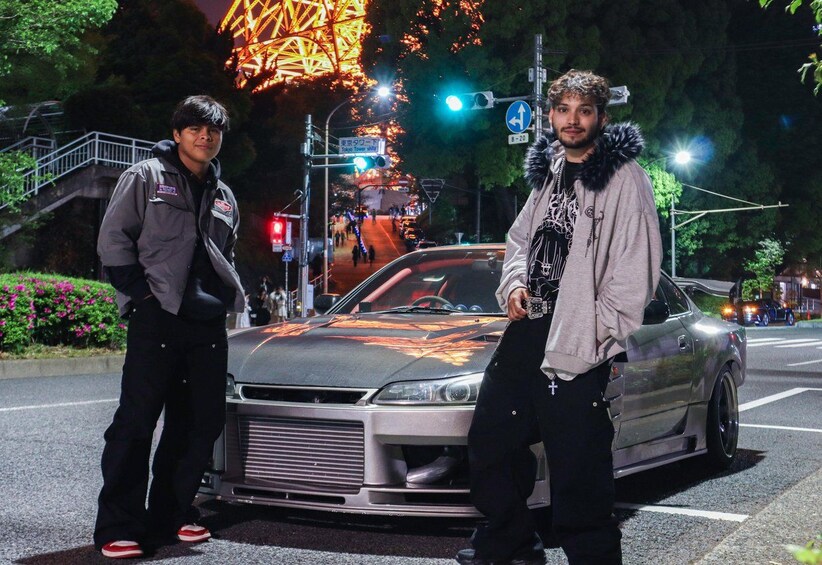 Two people standing beside a modified car on a street at night with a brightly lit Ferris wheel in the background. 3