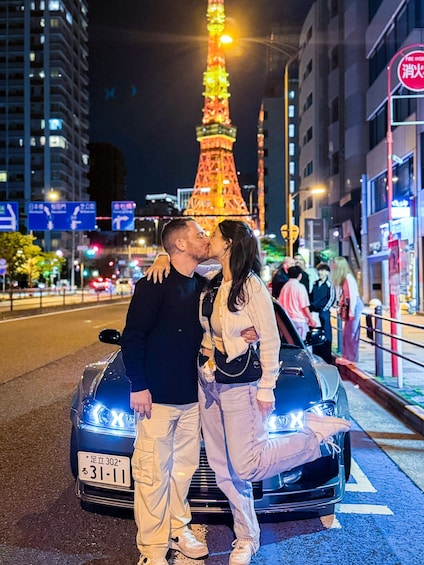 A couple kissing in front of a brightly lit car with a license plate, a tall illuminated tower in the background, and a busy urban street with buildings and signs. 1