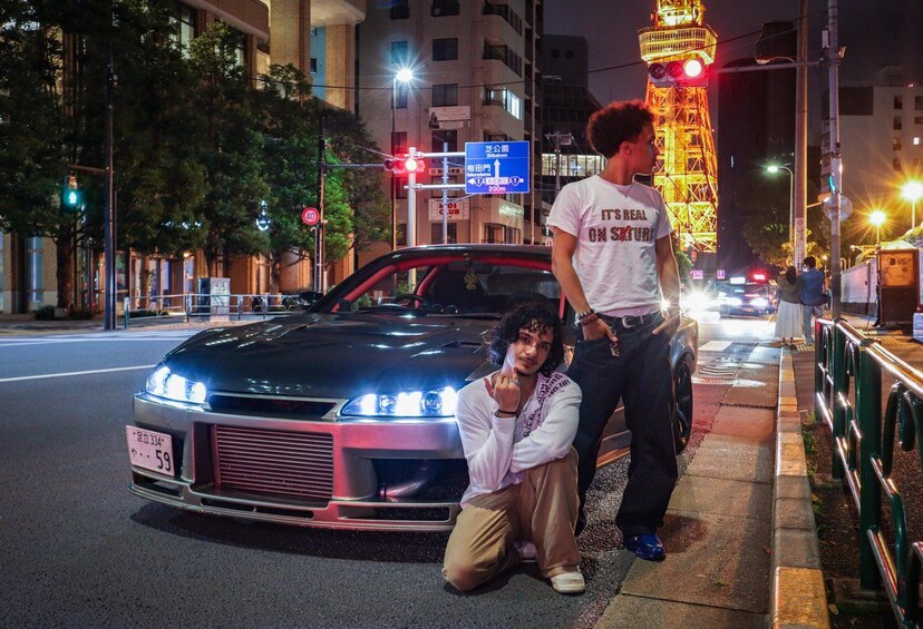 A nighttime urban scene with a brightly lit car, a person standing next to it, and a tower with illuminated lights in the background. 2