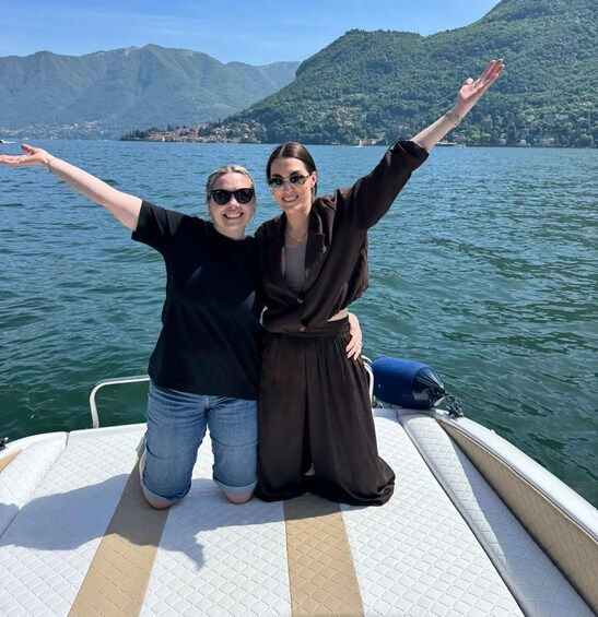 Two people on a boat with arms raised, a lake, and mountains in the background. 3