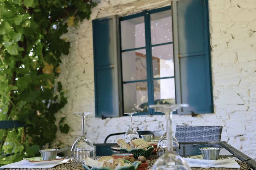 A table set outdoors with food and drinks, a window with blue shutters, and greenery in the background. 3