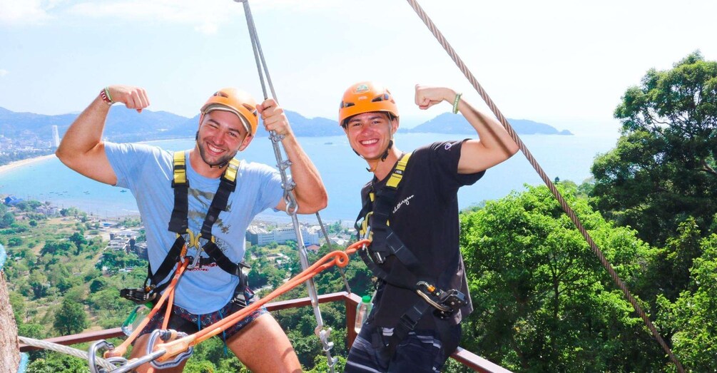 Two people on a zip line, wearing helmets and harnesses, with a scenic view of a coastline and greenery in the background. 1