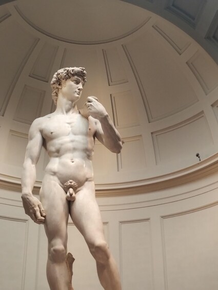 A marble statue of a muscular male figure in a classical pose, standing in a room with a domed ceiling and decorative moldings. 1