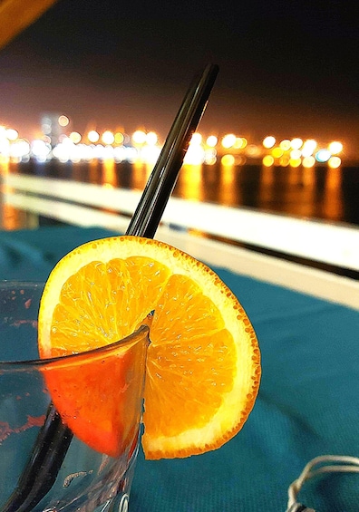 Cóctel con una rodaja de naranja y una pajita, sobre un fondo difuminado de paisaje urbano nocturno. 5