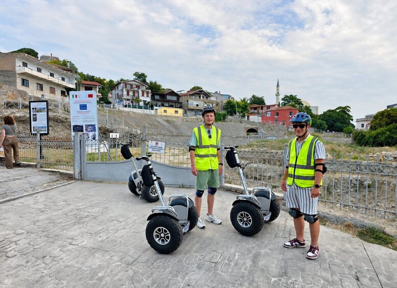 Dos personas con chalecos de seguridad de pie junto a patinetes Segway en una zona pavimentada, con un cartel y una valla. 4