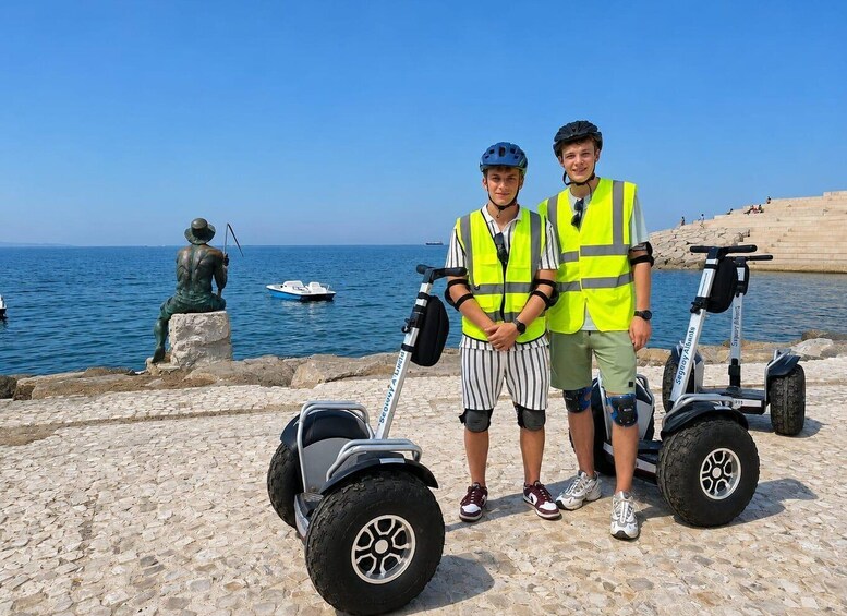 Dos personas sobre un pavimento de piedra junto al mar, usando chalecos reflectantes y cascos, con una estatua y un bote al fondo. 2