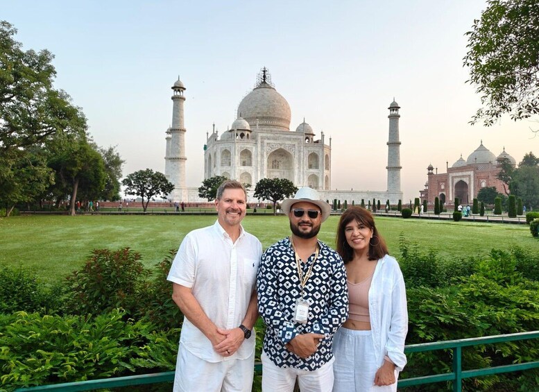 Drie personen staan voor de Taj Mahal, met een verzorgde tuin en een heldere lucht. 1