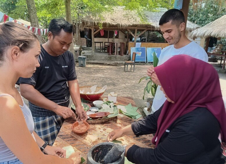 A group of people engaged in a traditional food preparation activity outdoors, with a thatched-roof structure and a patterned tablecloth in the background. 3