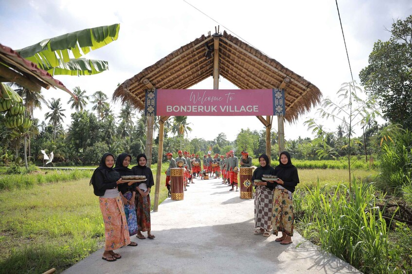 A village entrance with a sign reading 'Welcome to BONJERUK VILLAGE' under a thatched roof. 1