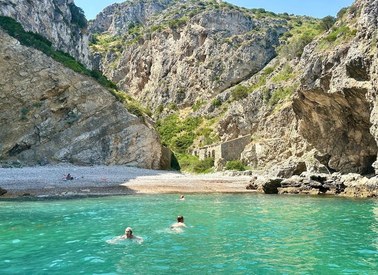 A rocky beach with clear turquoise water, people swimming, and a mountainous backdrop. 3