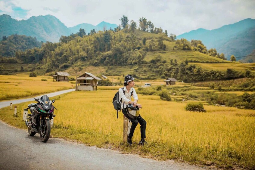 A motorcycle parked on a rural road with a person sitting on a post, surrounded by fields and mountains in the background. 1