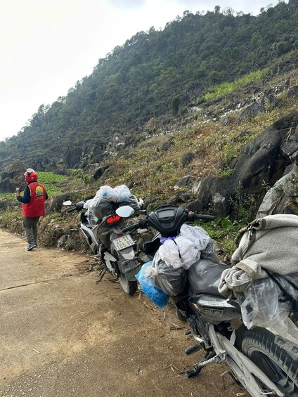 A person in a red jacket standing next to a motorcycle with a license plate, surrounded by other motorcycles and bags on a dirt path with a mountainous backdrop. 5
