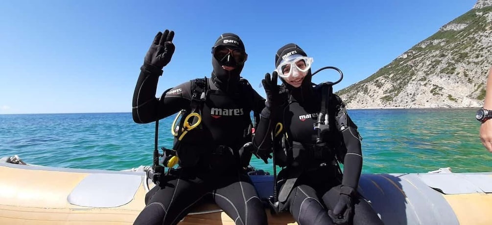 Two divers in black wetsuits with 'mares' branding on their gear, sitting on a boat with a clear blue sea and a rocky coastline in the background. 3