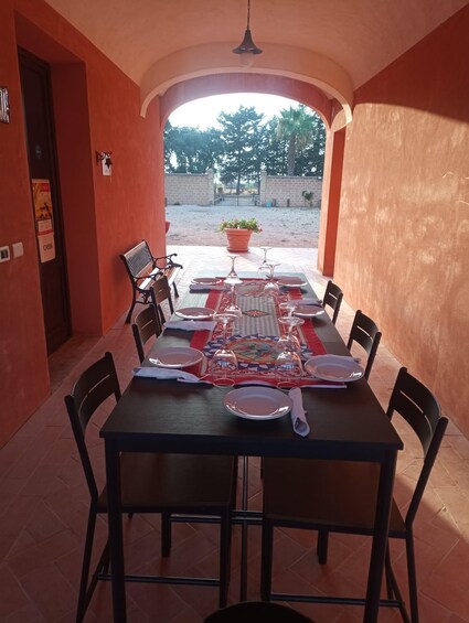 A dining table set with plates, glasses, and a placemat in a sunlit outdoor area with a view of trees and a gate. 1