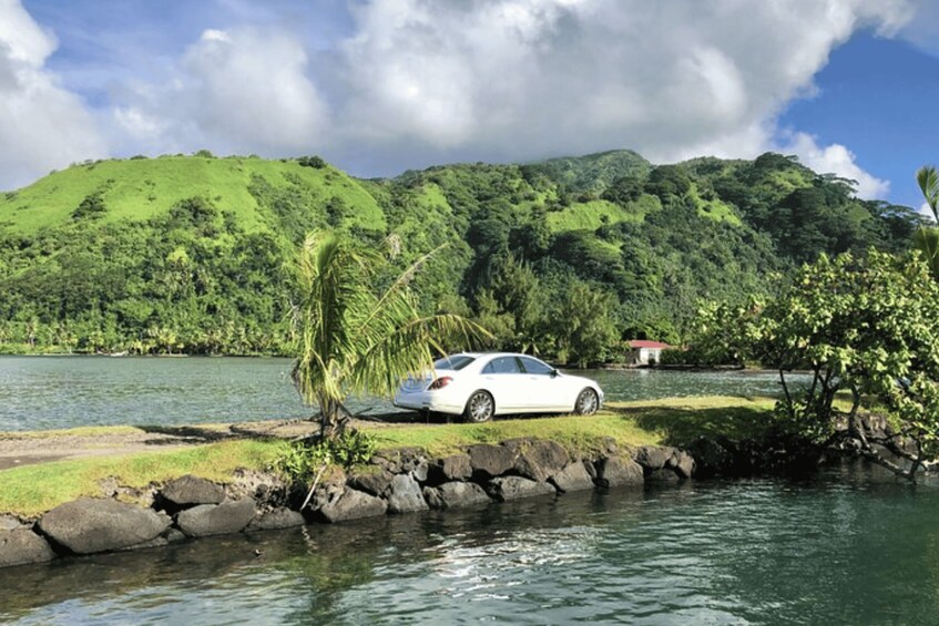 A white car parked by a calm lake with lush green hills in the background. 2