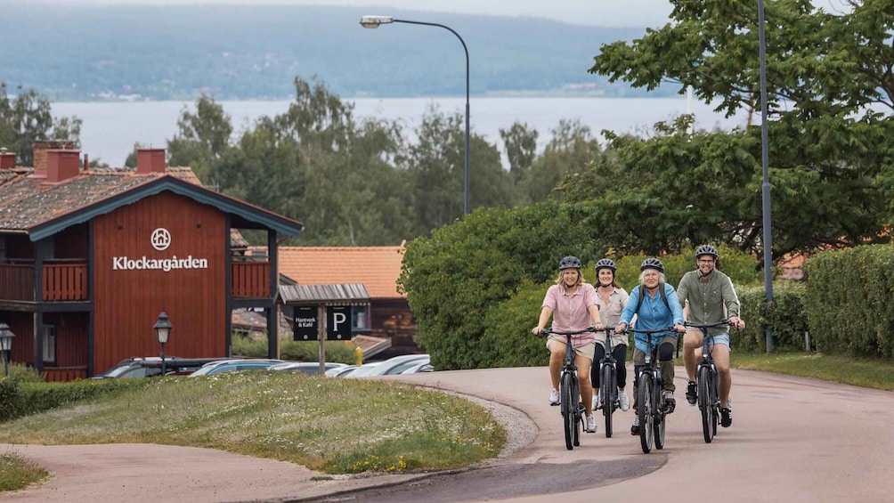 Four people riding bicycles on a paved path with a wooden building labeled 'Klockargarden' in the background. 3