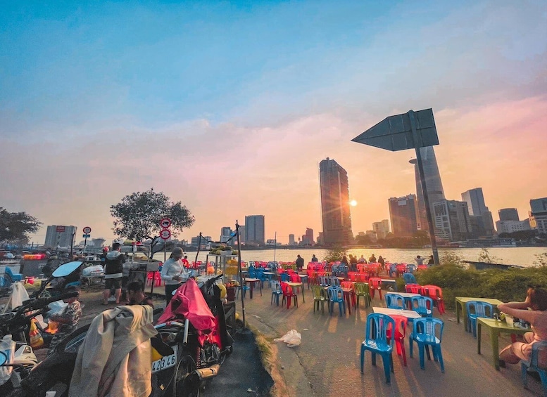 A riverside outdoor market with colorful plastic chairs and tables, a large umbrella, and a city skyline in the background during sunset. 1