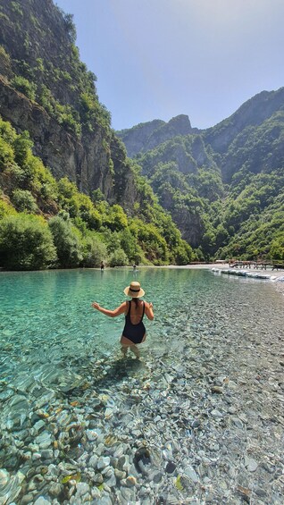 A person wading through clear water in a rocky riverbed with lush green mountains in the background. 1