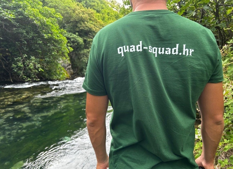 A person wearing a green t-shirt with 'quad-squad.hr' printed on the back, standing by a flowing river surrounded by lush greenery. 2