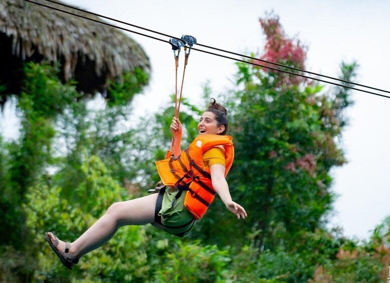 A person ziplining, wearing a life jacket and harness, with greenery and a thatched roof structure in the background. 5
