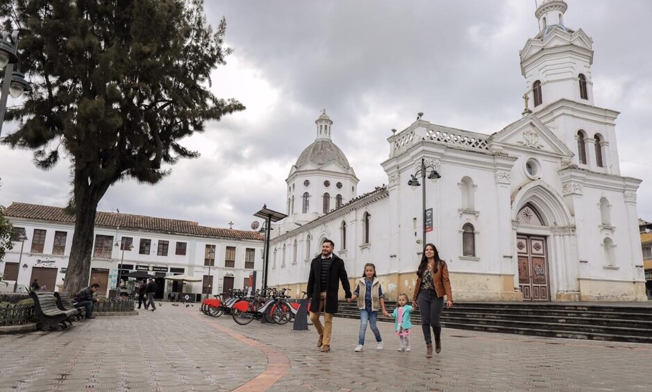 A cobblestone plaza with a large white church, a tree, and people walking. 2