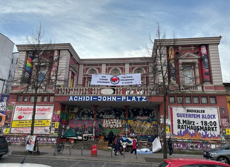 A building with graffiti and banners, including one for 'Achidi-John-Platz' and another for 'QueerFem Block'. 1