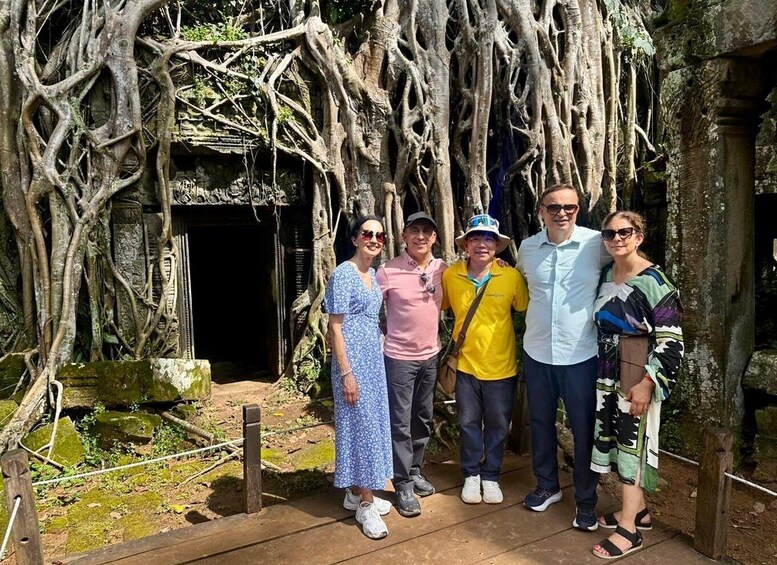 A group of people standing in front of a tree with extensive roots and a stone structure. 3