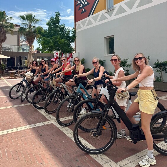 A group of people on stationary bikes in an outdoor setting with a brick pavement and a building with a geometric design in the background. 2