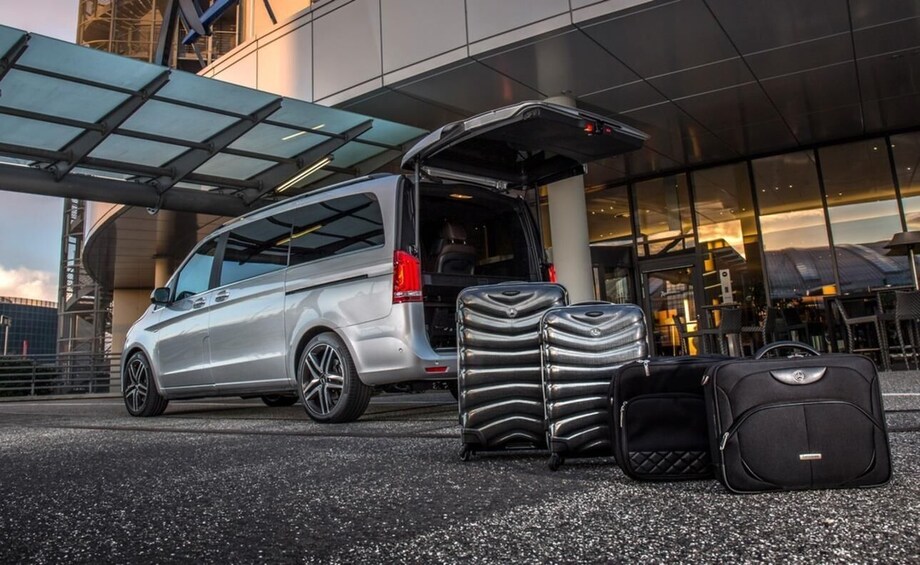 A silver van with its back door open, luggage, and a building with a glass facade in the background. 1