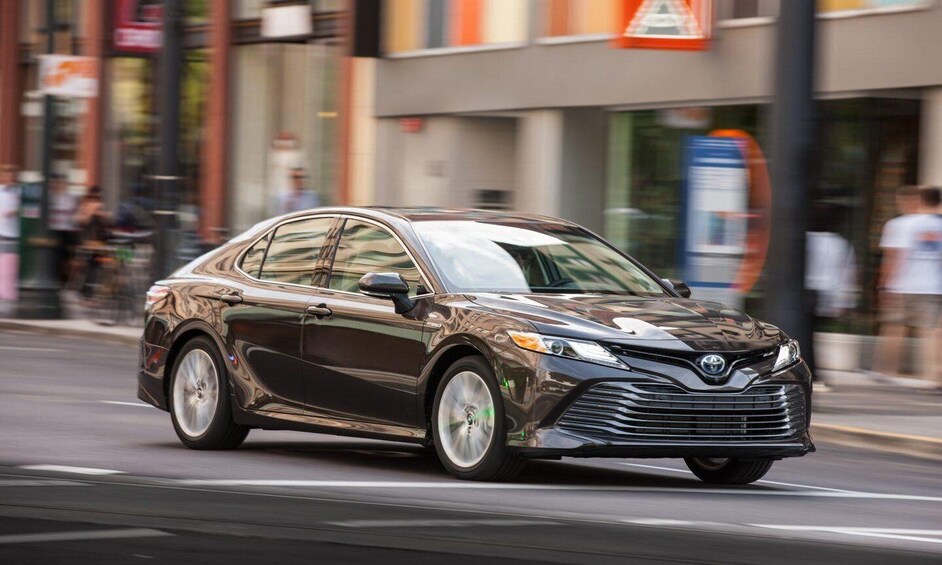 A dark-colored sedan driving on a city street with storefronts and pedestrians in the background. 3