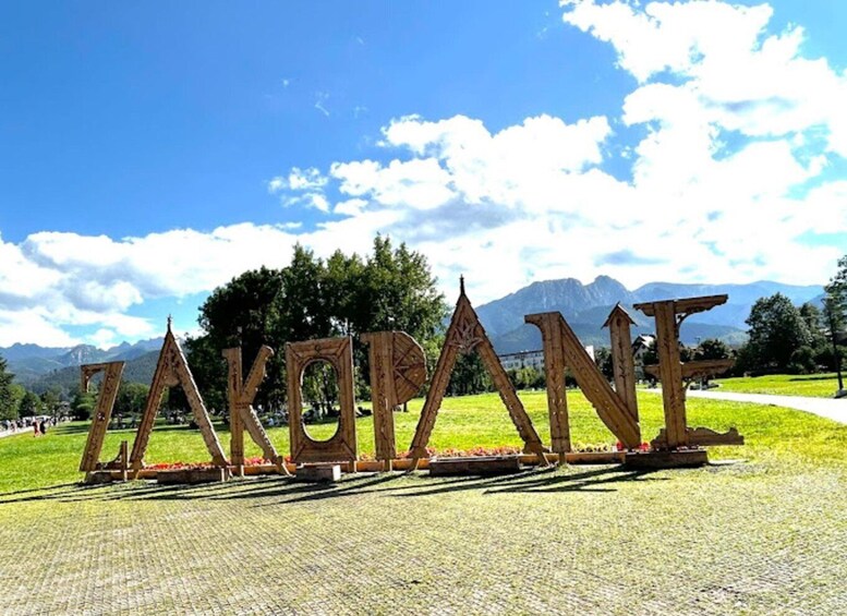 A wooden sign spelling 'ZAKOPANE' in a park with mountains in the background. 1