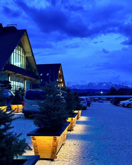 A building with a sloped roof and large windows, surrounded by parked cars and a gravel driveway. 2