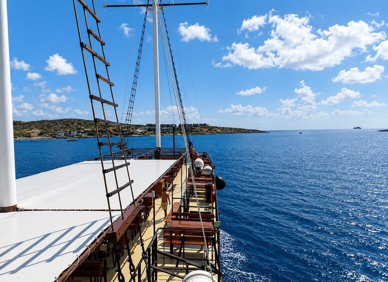 A ship's deck with a person walking, a white mast, and a coastline with buildings in the background. 3