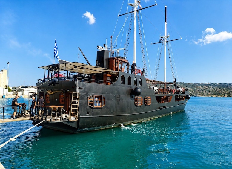 A large, dark-toned ship docked at a pier with clear blue water, a flag, and a hilly landscape in the background. 1