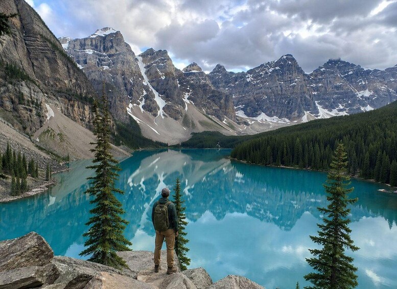 A person standing on a rocky outcrop overlooking a turquoise lake surrounded by mountains and dense forests. 2