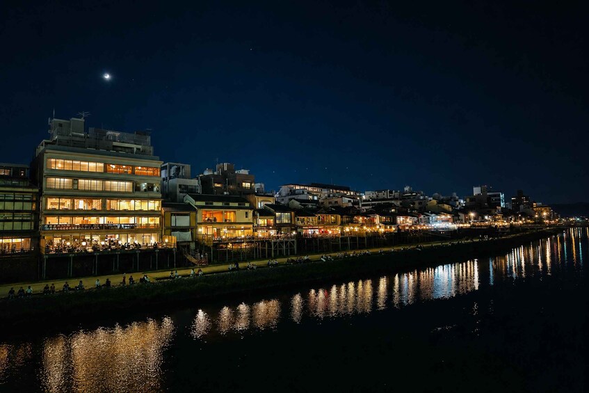 A nighttime cityscape with illuminated buildings along a river, a visible moon, and a reflection of lights on the water. 5