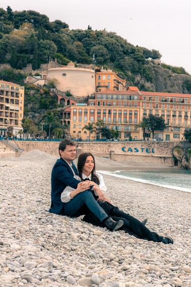 A couple sitting on a pebbled beach with a backdrop of buildings and a hill. 4