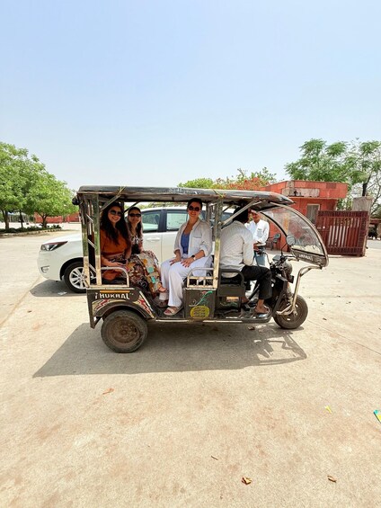 A tuk-tuk with four passengers, parked in a lot with trees and a building in the background. 5
