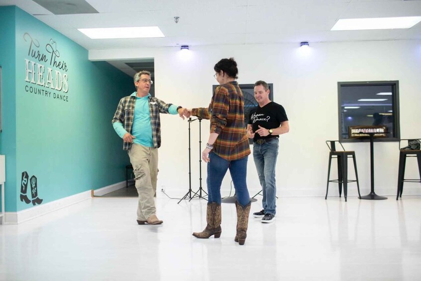 Three people in a room with teal walls and a sign that says 'Turn Their HEADS COUNTRY DANCE.' 3