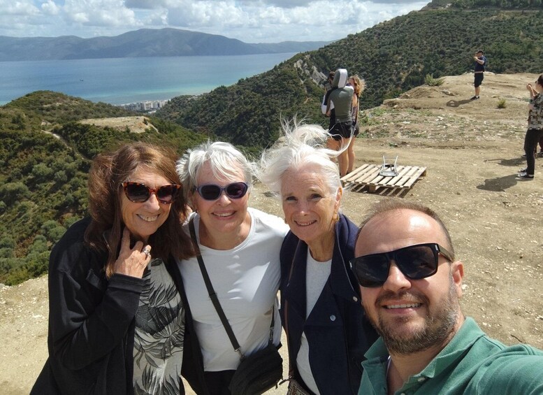 A group of people taking a selfie on a hillside with a view of a body of water and mountains in the background. 4
