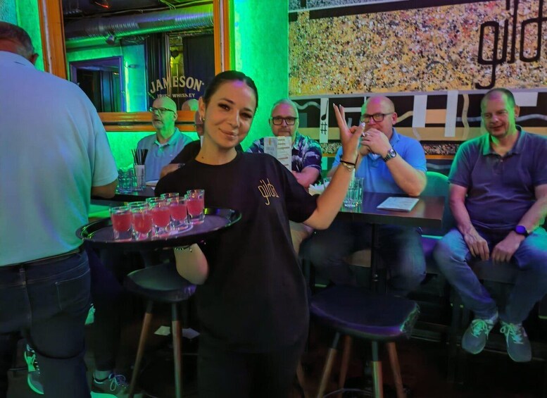 A person holding a tray with drinks in a bar with green lighting and a 'JAMESON' sign in the background. 2