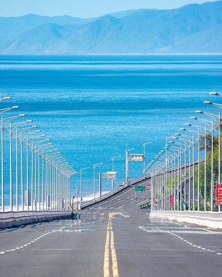 A coastal road with streetlights, a bridge, and a view of the ocean and mountains. 3