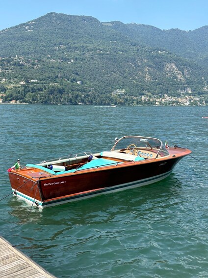 A wooden boat named 'The View Como Lake 2' docked at a pier with a mountainous landscape in the background. 2