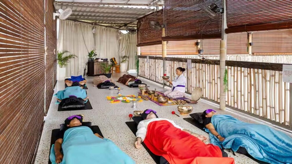 A group of people in a covered outdoor area, some lying on mats, others sitting, with traditional bowls and flowers on the floor. 3