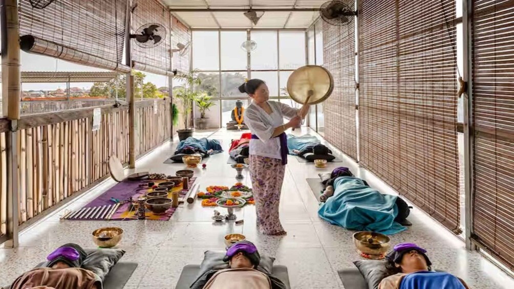 A person playing a drum in a room with several people lying on mats, surrounded by musical instruments and flowers. 4